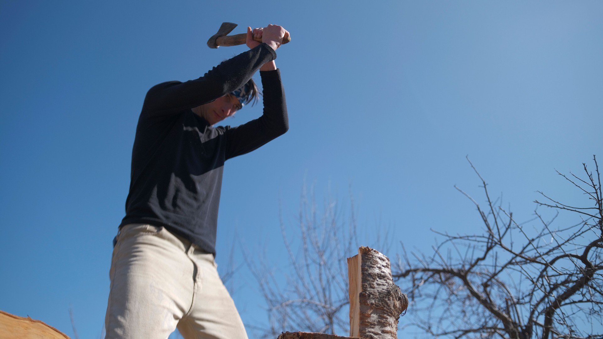 A white adult man is chopping firewood with an axe in the fresh air. The lumberjack. The sky is blue and the trees are bare. The man is focused on his task